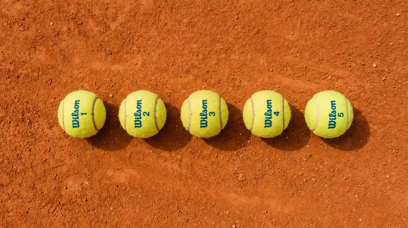 Mehrere Tennisbälle auf einem Sandplatz nebeneinander angeordnet als Symbol für kombinierte Tenniswetten